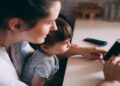 A parent using a smartphone while supervising a child, highlighting online safety and AI-related privacy risks.